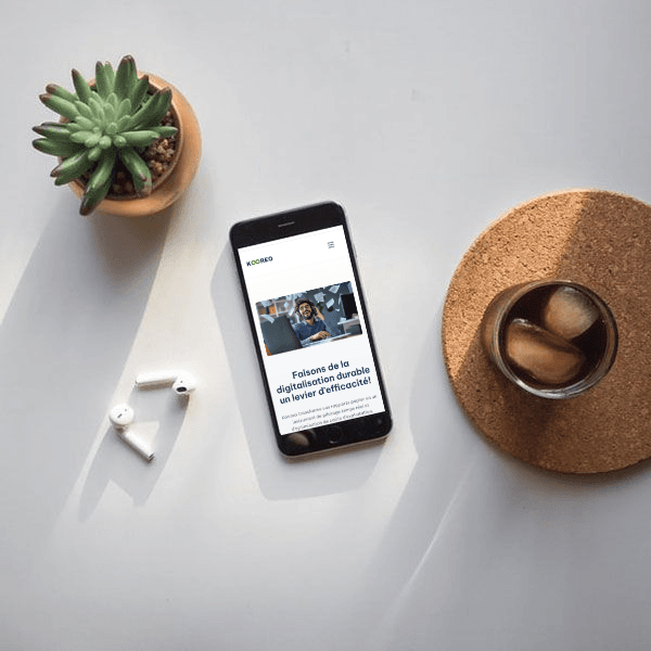 Un smartphone sur une table avec une plante, une boisson et des écouteurs. sur l'écran la page d'accueil du site web kooreo.fr 
